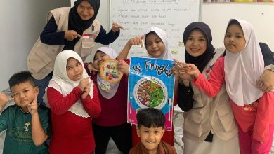 Mahasiswi KKN Internasional UMS bersama anak-anak perantauan di Malaysia memegang poster panduan porsi Isi Piringku Kemenkes di depan papan tulis 10 Pedoman Gizi Seimbang.