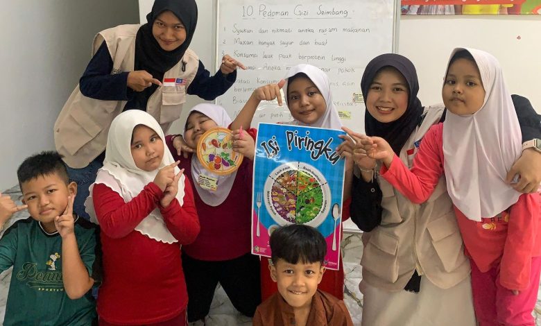Mahasiswi KKN Internasional UMS bersama anak-anak perantauan di Malaysia memegang poster panduan porsi Isi Piringku Kemenkes di depan papan tulis 10 Pedoman Gizi Seimbang.