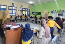 Foto suasana dalam ruang kelas di MIM Kauman saat momen pembagian rapor unik. Siswa, orang tua, dan guru terlihat duduk bersama di meja siswa, berinteraksi dan berpartisipasi dalam aktivitas parenting imersif.