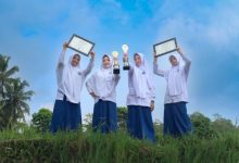 Empat santriwati MTs Almatera Temanggung berpose memegang dua piala dan dua sertifikat penghargaan TKA nasional di area persawahan di bawah langit biru.