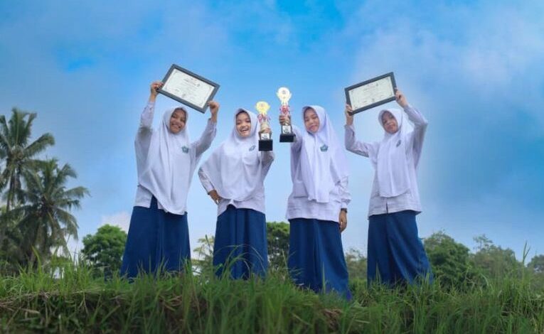 Empat santriwati MTs Almatera Temanggung berpose memegang dua piala dan dua sertifikat penghargaan TKA nasional di area persawahan di bawah langit biru.