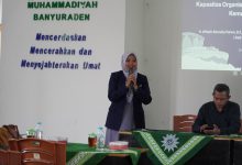 Seorang pembicara wanita berhijab sedang berbicara menggunakan mikrofon di acara Muhammadiyah Banyuraden bersama seorang pria di meja pembicara.