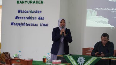 Seorang pembicara wanita berhijab sedang berbicara menggunakan mikrofon di acara Muhammadiyah Banyuraden bersama seorang pria di meja pembicara.