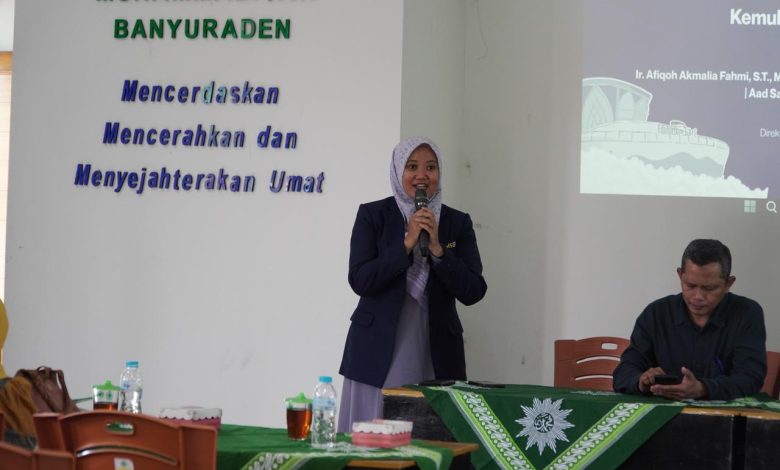 Seorang pembicara wanita berhijab sedang berbicara menggunakan mikrofon di acara Muhammadiyah Banyuraden bersama seorang pria di meja pembicara.