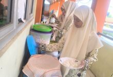 Dua peserta didik mengenakan jilbab dan seragam bermotif sedang mengambil makanan dalam suasana prasmanan.
