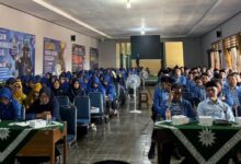 Peserta seminar yang terdiri dari bapak-bapak dan ibu-ibu dengan seragam biru sedang duduk rapi menyimak acara di dalam ruangan aula.