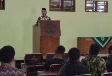 Seorang pria mengenakan peci dan baju batik sedang memberikan sambutan di atas mimbar di hadapan peserta rapat.