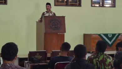 Seorang pria mengenakan peci dan baju batik sedang memberikan sambutan di atas mimbar di hadapan peserta rapat.