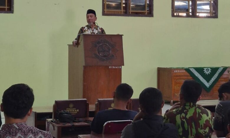 Seorang pria mengenakan peci dan baju batik sedang memberikan sambutan di atas mimbar di hadapan peserta rapat.