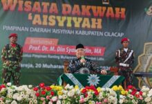 Prof. Dr. M. Din Syamsuddin, MA, memberikan sambutan di panggung Wisata Dakwah Aisyiyah PDA Cilacap, didampingi dua anggota KOKAM. Di depan panggung terdapat rangkaian bunga yang indah.