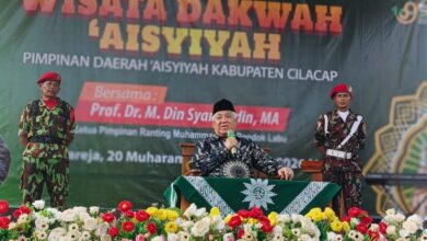 Prof. Dr. M. Din Syamsuddin, MA, memberikan sambutan di panggung Wisata Dakwah Aisyiyah PDA Cilacap, didampingi dua anggota KOKAM. Di depan panggung terdapat rangkaian bunga yang indah.