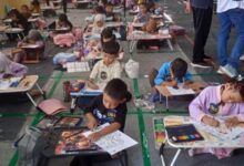 Sekelompok anak-anak, termasuk beberapa dengan jilbab dan beberapa tanpa, duduk di lantai dan di meja portabel kecil, berfokus mewarnai. Di latar belakang, ada lebih banyak anak dan orang dewasa yang berdiri, dan sebuah maskot.