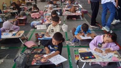 Sekelompok anak-anak, termasuk beberapa dengan jilbab dan beberapa tanpa, duduk di lantai dan di meja portabel kecil, berfokus mewarnai. Di latar belakang, ada lebih banyak anak dan orang dewasa yang berdiri, dan sebuah maskot.