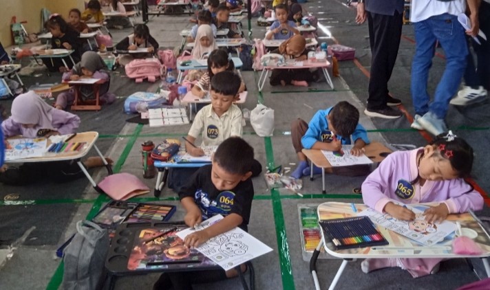 Sekelompok anak-anak, termasuk beberapa dengan jilbab dan beberapa tanpa, duduk di lantai dan di meja portabel kecil, berfokus mewarnai. Di latar belakang, ada lebih banyak anak dan orang dewasa yang berdiri, dan sebuah maskot.