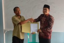 Pria berbaju batik (Ahmad Wildan, S.Sos.) berjabat tangan dengan pria berbaju hijau, menerima map dokumen, di depan papan tulis putih, pada acara serah terima jabatan kepala SMP Islam Pamotan yang tertulis di map.
