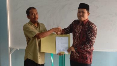 Pria berbaju batik (Ahmad Wildan, S.Sos.) berjabat tangan dengan pria berbaju hijau, menerima map dokumen, di depan papan tulis putih, pada acara serah terima jabatan kepala SMP Islam Pamotan yang tertulis di map.