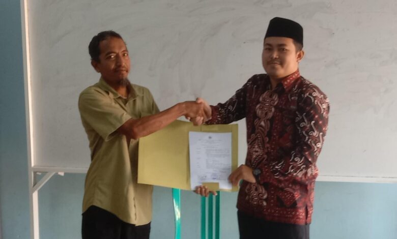 Pria berbaju batik (Ahmad Wildan, S.Sos.) berjabat tangan dengan pria berbaju hijau, menerima map dokumen, di depan papan tulis putih, pada acara serah terima jabatan kepala SMP Islam Pamotan yang tertulis di map.