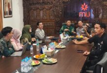 Sekelompok orang sedang melakukan pertemuan di ruang rapat yang dihiasi ukiran kayu tradisional dan logo Lazismu, dengan hidangan buah dan botol air di meja.Judul: Pertemuan Strategis Lazismu di Ruang Rapat Ukiran Kayu