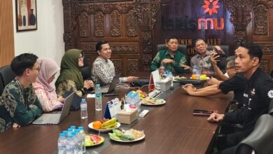 Sekelompok orang sedang melakukan pertemuan di ruang rapat yang dihiasi ukiran kayu tradisional dan logo Lazismu, dengan hidangan buah dan botol air di meja.Judul: Pertemuan Strategis Lazismu di Ruang Rapat Ukiran Kayu