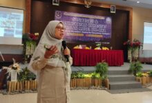 Wanita berhijab sedang berbicara di depan panggung dengan latar belakang spanduk bertuliskan "SD Muhammadiyah 1 Ketelan Surakarta" dan dua layar proyeksi menampilkan presentasi.
