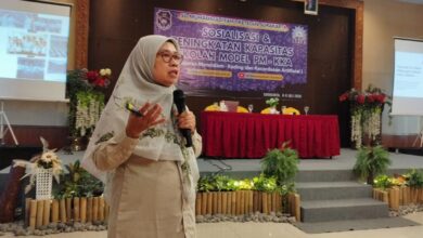 Wanita berhijab sedang berbicara di depan panggung dengan latar belakang spanduk bertuliskan "SD Muhammadiyah 1 Ketelan Surakarta" dan dua layar proyeksi menampilkan presentasi.