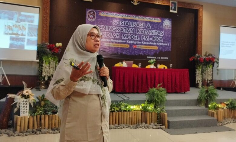 Wanita berhijab sedang berbicara di depan panggung dengan latar belakang spanduk bertuliskan "SD Muhammadiyah 1 Ketelan Surakarta" dan dua layar proyeksi menampilkan presentasi.