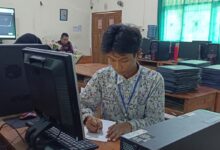 Siswa laki-laki dengan seragam batik sedang fokus menulis di meja depan komputer di lab komputer sekolah, dikelilingi oleh stasiun komputer lain dan latar belakang ruang kelas.