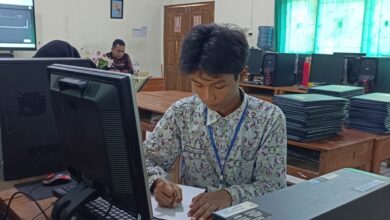 Siswa laki-laki dengan seragam batik sedang fokus menulis di meja depan komputer di lab komputer sekolah, dikelilingi oleh stasiun komputer lain dan latar belakang ruang kelas.