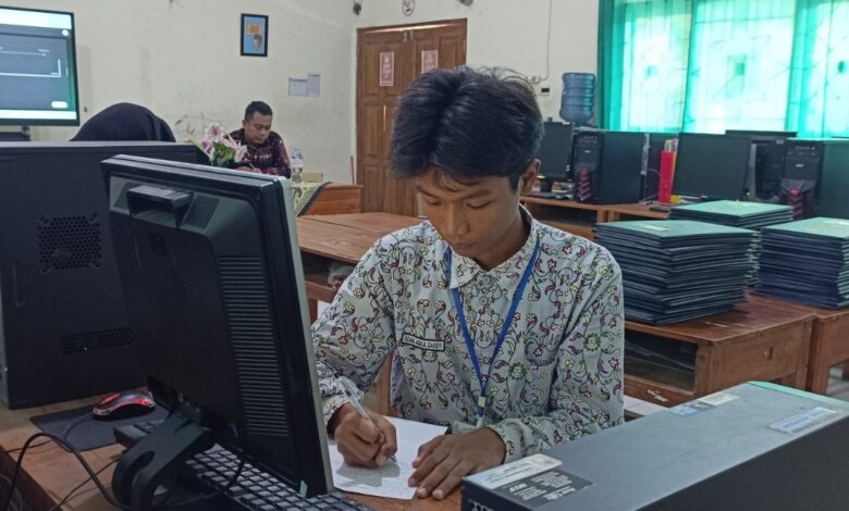 Siswa laki-laki dengan seragam batik sedang fokus menulis di meja depan komputer di lab komputer sekolah, dikelilingi oleh stasiun komputer lain dan latar belakang ruang kelas.