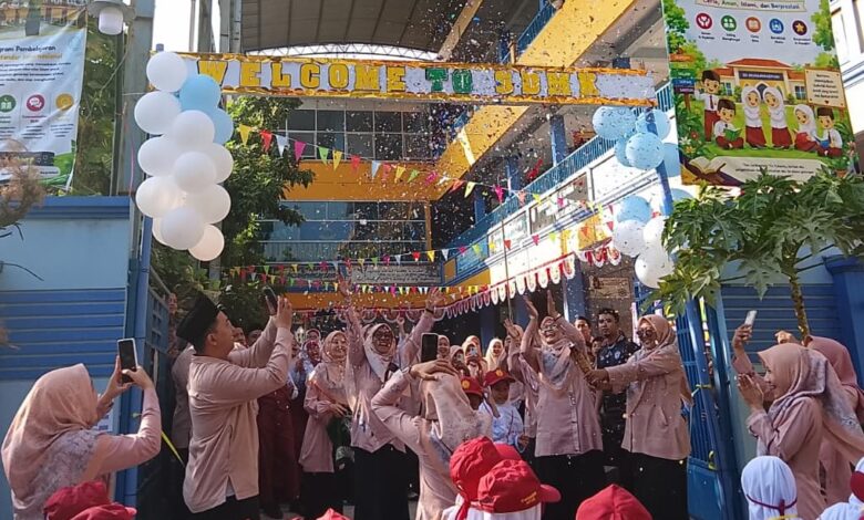 Suasana meriah pembukaan MPLS SD Muhammadiyah Kriyan 2026. Para guru dan orang tua bersorak melempar kertas konfeti ke udara di bawah spanduk besar 'Sekolah Ramah Anak' dan 'International Class Program (ICP)' serta balon putih biru di gerbang sekolah, disaksikan oleh murid baru bertopi merah.