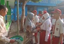 Seorang guru berhijab memberikan medali kepada siswi SD saat upacara sekolah, disaksikan siswa-siswa lain dan seorang guru pria di halaman sekolah dengan koridor hijau dan bangunan biru di background.