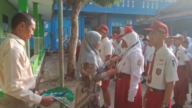 Seorang guru berhijab memberikan medali kepada siswi SD saat upacara sekolah, disaksikan siswa-siswa lain dan seorang guru pria di halaman sekolah dengan koridor hijau dan bangunan biru di background.