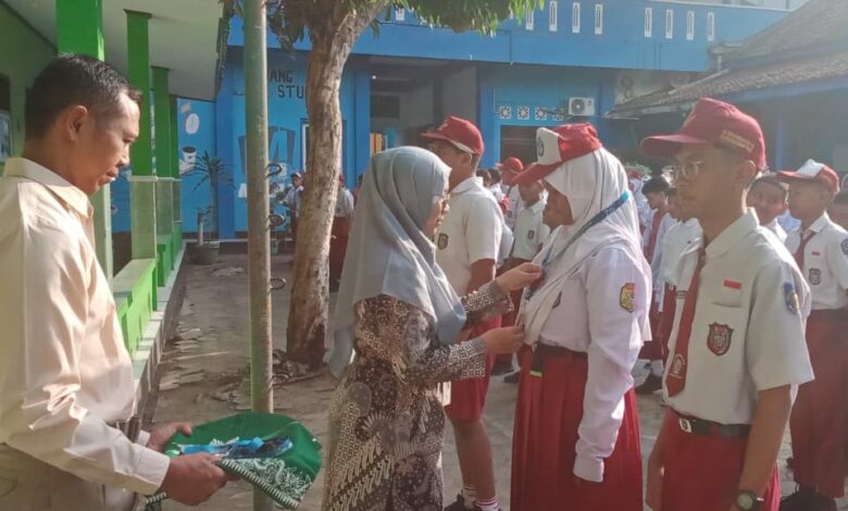 Seorang guru berhijab memberikan medali kepada siswi SD saat upacara sekolah, disaksikan siswa-siswa lain dan seorang guru pria di halaman sekolah dengan koridor hijau dan bangunan biru di background.