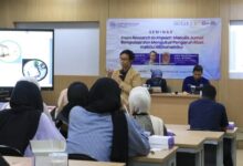 Sriyanto sedang memberikan materi seminar di depan peserta, fokus pada cara menulis artikel ilmiah standar ScienceDirect, dengan spanduk seminar "From Research to Impact: Menulis Jurnal Bereputasi dan Mengukur Pengaruh Riset melalui Bibliometrika" di latar belakang.