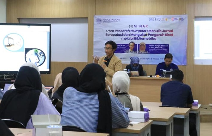 Sriyanto sedang memberikan materi seminar di depan peserta, fokus pada cara menulis artikel ilmiah standar ScienceDirect, dengan spanduk seminar "From Research to Impact: Menulis Jurnal Bereputasi dan Mengukur Pengaruh Riset melalui Bibliometrika" di latar belakang.