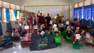 Sekelompok anak sekolah dasar berseragam hijau di dalam kelas sedang berpose bersama anggota BikersMu, menunjukkan buku-buku baru yang mereka terima sebagai bagian dari aksi sosial. Di bagian depan tengah, terdapat bendera atau spanduk hitam bertuliskan "BIKERS MU".