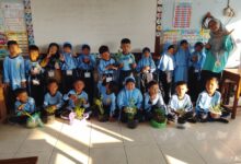 Guru dan murid sekolah dasar berpose bersama sambil memegang tanaman dalam pot di dalam ruang kelas dengan dekorasi edukatif di dinding.