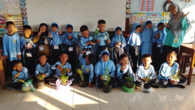 Guru dan murid sekolah dasar berpose bersama sambil memegang tanaman dalam pot di dalam ruang kelas dengan dekorasi edukatif di dinding.