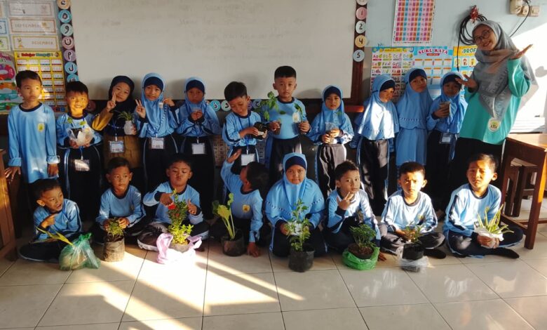 Guru dan murid sekolah dasar berpose bersama sambil memegang tanaman dalam pot di dalam ruang kelas dengan dekorasi edukatif di dinding.