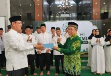 Seorang pria memberikan dokumen kepada pria lain dalam prosesi pelantikan resmi di depan jajaran pengurus organisasi dengan latar belakang spanduk acara.