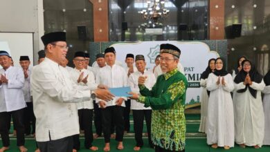Seorang pria memberikan dokumen kepada pria lain dalam prosesi pelantikan resmi di depan jajaran pengurus organisasi dengan latar belakang spanduk acara.