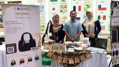 Tim mahasiswa berpose di stan pameran produk tas lipat "Arterra" dengan spanduk promosi dan berbagai contoh produk di meja.