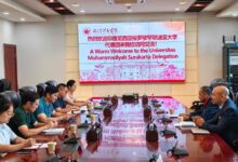 Sebuah pertemuan delegasi di ruang konferensi dengan meja kayu panjang dan layar proyeksi besar. Layar proyeksi besar di bagian depan menampilkan logo Wuhan Sports University dan teks sambutan berwarna merah: "A Warm Welcome to the Universitas Muhammadiyah Surakarta Delegation." Dua kelompok perwakilan, satu mengenakan kemeja seragam hijau dan putih dan satu lagi mengenakan jas, duduk berhadapan.
