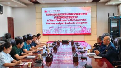 Sebuah pertemuan delegasi di ruang konferensi dengan meja kayu panjang dan layar proyeksi besar. Layar proyeksi besar di bagian depan menampilkan logo Wuhan Sports University dan teks sambutan berwarna merah: "A Warm Welcome to the Universitas Muhammadiyah Surakarta Delegation." Dua kelompok perwakilan, satu mengenakan kemeja seragam hijau dan putih dan satu lagi mengenakan jas, duduk berhadapan.
