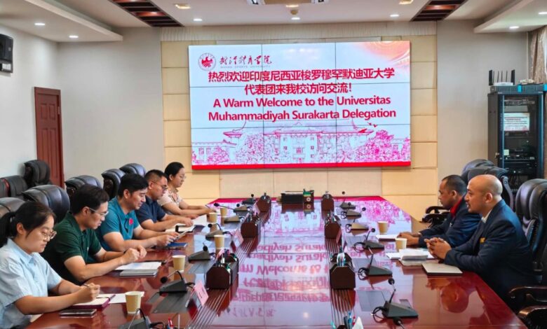 Sebuah pertemuan delegasi di ruang konferensi dengan meja kayu panjang dan layar proyeksi besar. Layar proyeksi besar di bagian depan menampilkan logo Wuhan Sports University dan teks sambutan berwarna merah: "A Warm Welcome to the Universitas Muhammadiyah Surakarta Delegation." Dua kelompok perwakilan, satu mengenakan kemeja seragam hijau dan putih dan satu lagi mengenakan jas, duduk berhadapan.