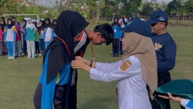 Seorang pejabat wanita sedang menyematkan tanda pengenal kepada peserta pelatihan disiplin di tengah lapangan.