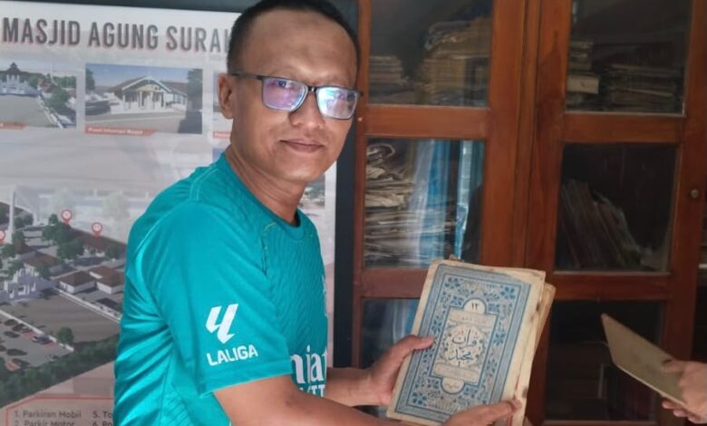Seorang pria berkacamata mengenakan kaos hijau menunjukkan kitab Al-Qur'an kuno di kompleks Masjid Agung Surakarta.
