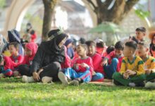Seorang guru berhijab hitam berinteraksi dengan ceria bersama sekelompok anak-anak berseragam sekolah yang duduk di atas rumput taman.