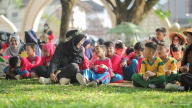 Seorang guru berhijab hitam berinteraksi dengan ceria bersama sekelompok anak-anak berseragam sekolah yang duduk di atas rumput taman.