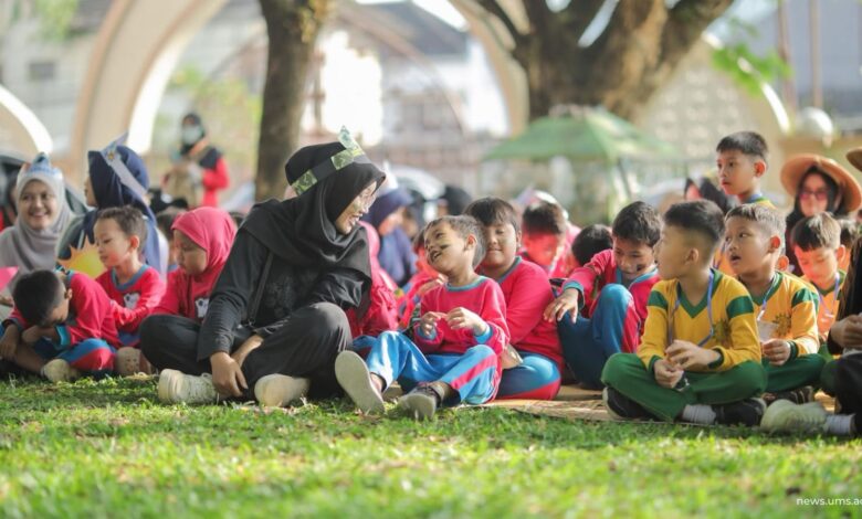 Seorang guru berhijab hitam berinteraksi dengan ceria bersama sekelompok anak-anak berseragam sekolah yang duduk di atas rumput taman.